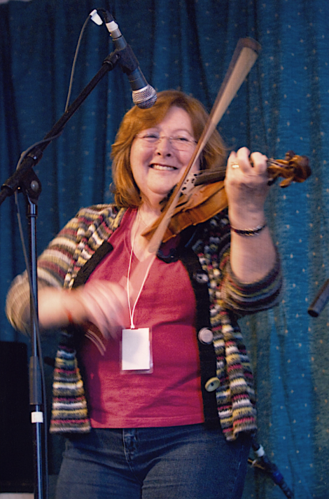 A person with short hair is playing the violin on stage, wearing a striped cardigan and a pink shirt, with a microphone positioned nearby.