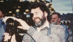 A man with a beard and patterned sweater is using a large video camera outdoors. Another person is visible in the background.