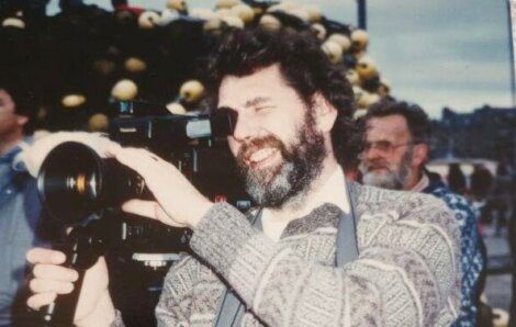 A man with a beard and patterned sweater is using a large video camera outdoors. Another person is visible in the background.