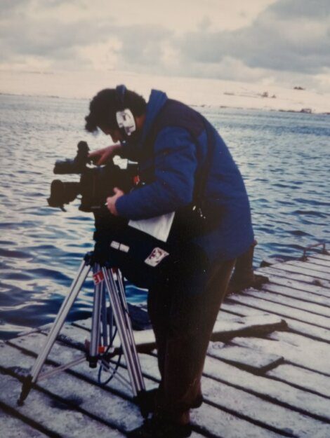 A person wearing a blue jacket and headphones operates a camera on a tripod, positioned on a wooden dock by a body of water with a snowy landscape in the background.