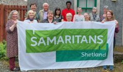 A diverse group of people standing outdoors holding a large banner that reads "SAMARITANS Shetland." They are in front of a gray building with a red fence on the left.