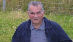 A man with gray hair and a blue jacket poses outdoors in front of a grass field and wire fence.