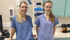 Two healthcare professionals are standing in a clinical setting, wearing scrubs and smiling at the camera. Medical equipment and supplies are visible in the background.