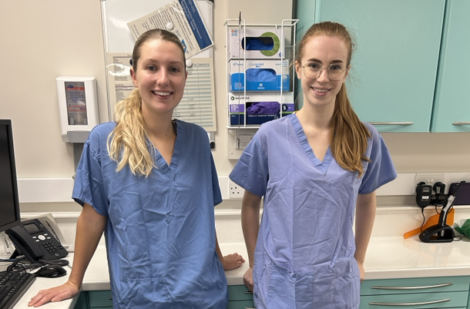 Two healthcare professionals are standing in a clinical setting, wearing scrubs and smiling at the camera. Medical equipment and supplies are visible in the background.