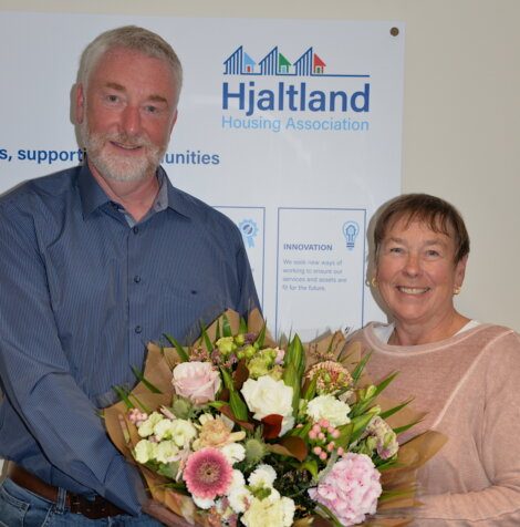 Two people standing in front of a Hjaltland Housing Association sign, smiling and holding a bouquet of flowers.