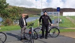 Two men stand beside parked bicycles on a paved path near a body of water and buildings. One man shields his eyes from the sun while the other wears a helmet and leans on his bike.