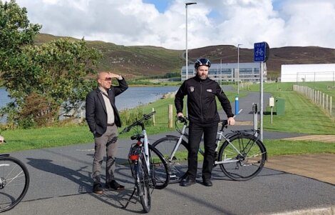 Two men stand beside parked bicycles on a paved path near a body of water and buildings. One man shields his eyes from the sun while the other wears a helmet and leans on his bike.