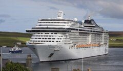 A large white cruise ship is docked at a port with green hills in the background and a smaller ferry nearby.