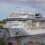 A large white cruise ship is docked at a port with green hills in the background and a smaller ferry nearby.