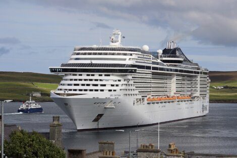 A large white cruise ship is docked at a port with green hills in the background and a smaller ferry nearby.