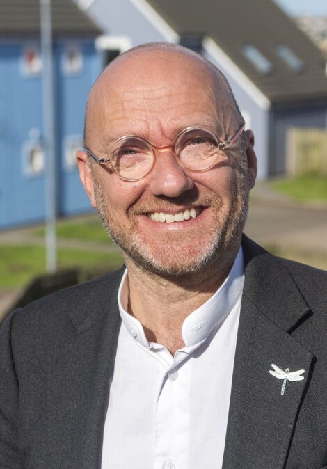 A smiling bald man with glasses and a beard wears a dark blazer and white shirt, with a dragonfly pin on his lapel. Background shows a blue building and green grass.