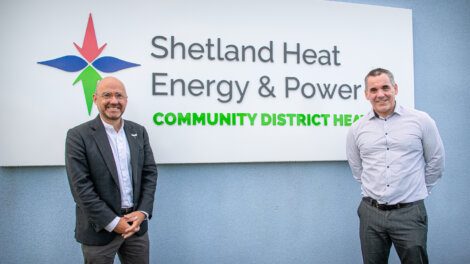 Two men stand in front of a sign reading "Shetland Heat Energy & Power Community District Heat." Both are smiling, one wearing a suit jacket and the other in a button-down shirt.