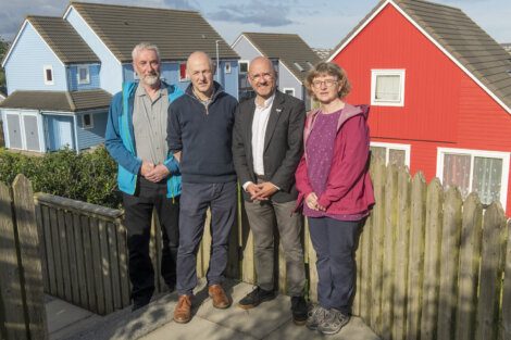 Four people stand side by side in front of colorful houses with a wooden fence in the foreground.