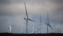 A group of large wind turbines stands on a grassy hill under a cloudy sky.