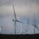 A group of large wind turbines stands on a grassy hill under a cloudy sky.