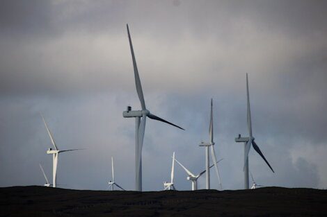 A group of large wind turbines stands on a grassy hill under a cloudy sky.