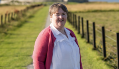 A smiling individual stands on a path flanked by fields and fences, wearing a white shirt and a pink cardigan.