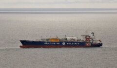 A ship with "INEOS" and "BELSTAFF" logos, along with "#BUILT FOR LIFE," sails on the ocean under a cloudy sky.