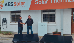 Two men shaking hands outside the AES Solar office with solar panels and a Tesla Powerwall visible. The building features signage for AES Solar and Solar Thermal.