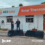 Two men shaking hands outside the AES Solar office with solar panels and a Tesla Powerwall visible. The building features signage for AES Solar and Solar Thermal.