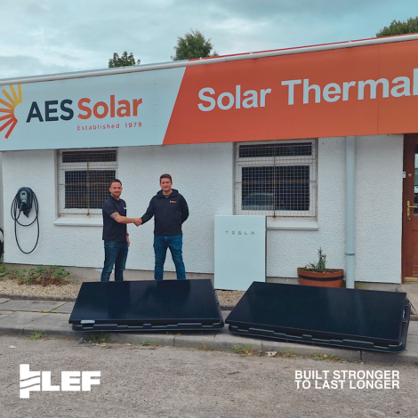 Two men shaking hands outside the AES Solar office with solar panels and a Tesla Powerwall visible. The building features signage for AES Solar and Solar Thermal.