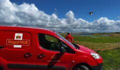 A Royal Mail van is parked on grass next to a person in an orange jacket. A drone is flying above the field with a partly cloudy sky in the background.