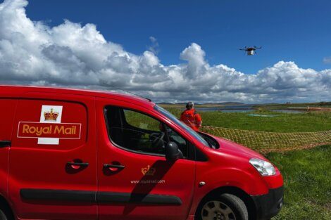 A Royal Mail van is parked on grass next to a person in an orange jacket. A drone is flying above the field with a partly cloudy sky in the background.