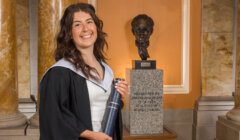 A woman in a graduation gown holds a diploma, standing in front of a pedestal with a bust statue and an inscribed plaque.