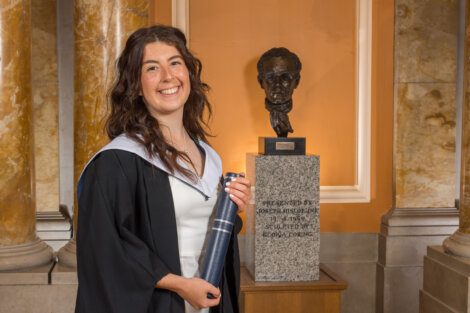 A woman in a graduation gown holds a diploma, standing in front of a pedestal with a bust statue and an inscribed plaque.