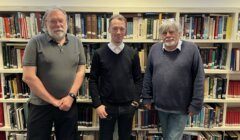 Three men are standing in front of a filled bookcase. The man in the middle wears a black sweater over a shirt, while the men on the sides wear casual shirts and sweaters.