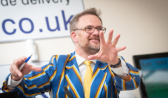 A man in a colorful striped jacket and yellow tie gestures with his hand while holding a pen, standing indoors. A wall sign and monitor are visible in the background.