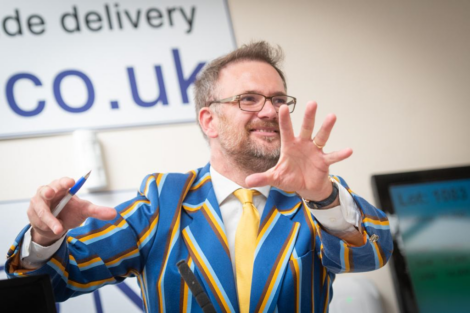 A man in a colorful striped jacket and yellow tie gestures with his hand while holding a pen, standing indoors. A wall sign and monitor are visible in the background.