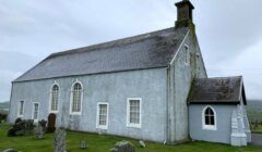 A gray stone church with arched windows and a small side entrance stands amid a grassy cemetery on an overcast day.