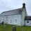 A gray stone church with arched windows and a small side entrance stands amid a grassy cemetery on an overcast day.