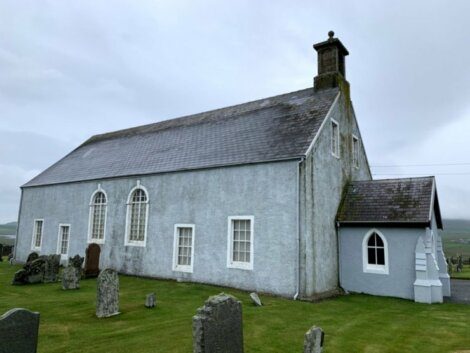 A gray stone church with arched windows and a small side entrance stands amid a grassy cemetery on an overcast day.