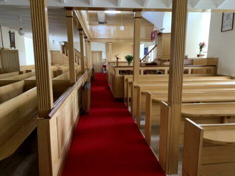 Interior of an empty church with wooden pews and columns, a red carpet running down the center aisle, and a pulpit area at the front.