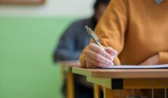 A person in an orange sweater is writing with a pen on paper at a desk in a classroom. Another person is blurred in the background.