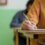 A person in an orange sweater is writing with a pen on paper at a desk in a classroom. Another person is blurred in the background.