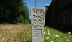 A stone tablet with intricate carvings stands on a grassy patch, featuring a circular design at the top and three human-like figures below.