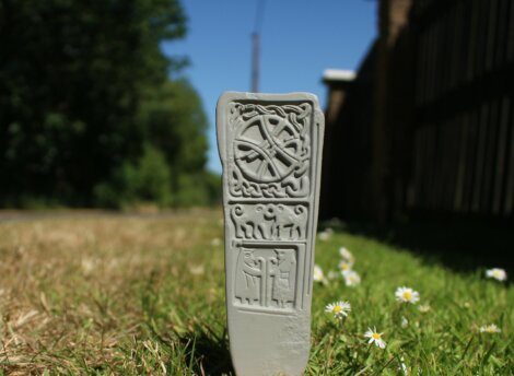 A stone tablet with intricate carvings stands on a grassy patch, featuring a circular design at the top and three human-like figures below.