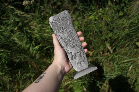 A hand holding a rectangular stone artifact featuring carved animal figures and an intricate circular design, with tall grass in the background.