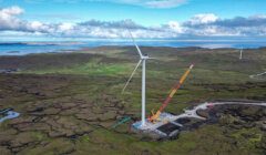 A large wind turbine under construction in a vast green and brown landscape, with a crane and several vehicles positioned at its base. Hills and water are visible in the background under a partly cloudy sky.