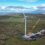 A large wind turbine under construction in a vast green and brown landscape, with a crane and several vehicles positioned at its base. Hills and water are visible in the background under a partly cloudy sky.