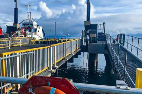 A docked ferry at a pier with yellow railings under a cloudy sky. The pier features walkways, equipment, and a lifebuoy in the foreground.