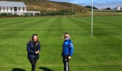 Two people stand on a neatly mowed sports field with a building and hills in the background.