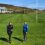 Two people stand on a neatly mowed sports field with a building and hills in the background.