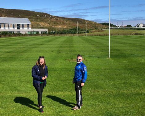 Two people stand on a neatly mowed sports field with a building and hills in the background.