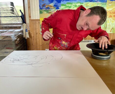 A person in a red jacket is creating abstract art on a white canvas with curved lines, using a speaker. Colorful paintings are in the background.