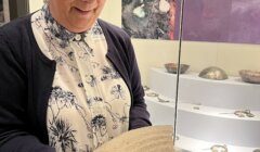 A person with glasses and gloves examines a large, ancient artifact in a museum display area. Exhibits and information panels are visible in the background.