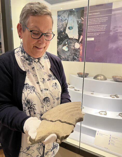 A person with glasses and gloves examines a large, ancient artifact in a museum display area. Exhibits and information panels are visible in the background.